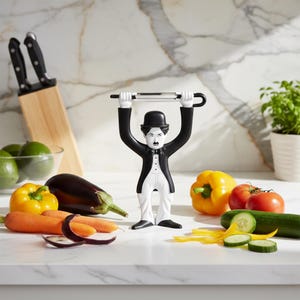 May include: A black and white Chaplin-themed vegetable peeler with a stainless steel blade, posed with fresh vegetables like carrots, tomatoes, and cucumbers. A wooden knife block with black knives is in the background.