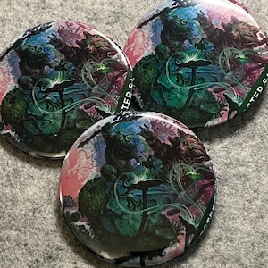 May include: Three circular pin-back buttons featuring a detailed fantasy illustration of a dark creature with glowing green accents, set against a vibrant pink and blue background. The artwork includes swirling lines and organic shapes.