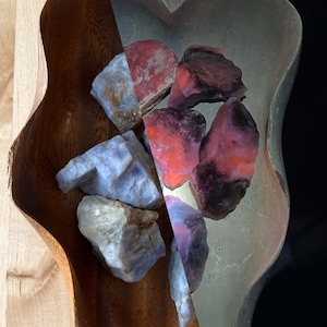 May include: A wooden bowl with a unique shape, filled with rough-cut gemstones in shades of red, purple, and blue. The bowl sits on a light-colored wooden surface, with a dark background. The gemstones are the focal point of the image.
