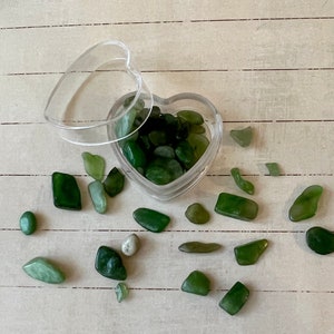 May include: A heart-shaped container filled with small, green, smooth stones. The container is open and some of the stones have spilled out onto a light wood surface.