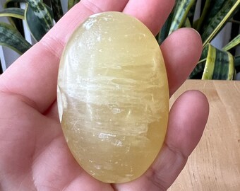 May include: A smooth, oval-shaped yellow gemstone with white streaks. The stone is held in a hand.