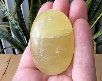 May include: A smooth, oval-shaped yellow calcite stone. The stone is held in a hand, showcasing its translucent quality and natural color variations. The stone is approximately 2 inches long, perfect for meditation or display.