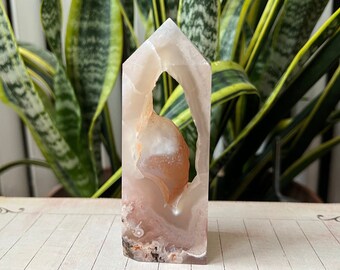 May include: A light pink and white agate crystal with a natural hole in the center, carved into a tower shape. The crystal is sitting on a lined paper surface.