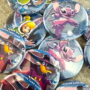 May include: A collection of circular pin-back buttons featuring various animated characters, including a blonde fairy in a green hood, a pink alien creature, and a purple duck in a hat, with the text TRADING CARD GAME visible on several items.