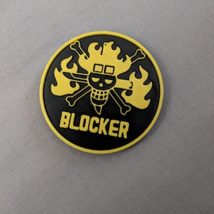 May include: A circular patch featuring a black background with a yellow border. The design displays a yellow skull and crossbones emblem surrounded by stylized flames, with the word BLOCKER printed in bold yellow capital letters below it.