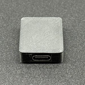 May include: A small, rectangular black device with a textured, grey top. The device has a USB-C port on one side. The item is likely an electronic component or adapter.