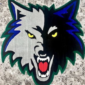 Pode incluir: Um tapete em forma de cabeça de lobo, dividido verticalmente em duas metades. Um lado é cinza, o outro preto. O lobo tem olhos amarelos, boca vermelha e dentes brancos. O tapete é contornado em verde e azul.