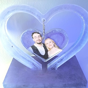 Può includere: Una cornice a forma di cuore nei toni del viola e del blu. La cornice contiene una fotografia di una coppia. La cornice è realizzata in un materiale traslucido e poggia su una base rettangolare.