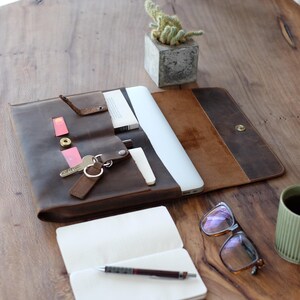 May include: Open brown leather laptop case with a laptop inside, keys, and cards. A small notebook with a pen, glasses, and a small cactus in a concrete pot are on a wooden table.