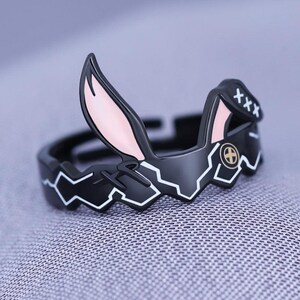May include: A black ring with a bunny ear design. The ring has a black base with white geometric accents and pink inner ear details. The ring features the text "XXX" on the side.