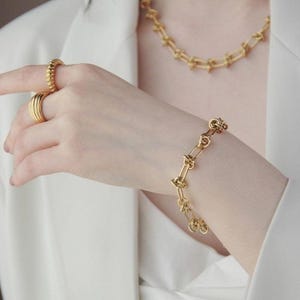Può includere: Un set di gioielli color oro con bracciale, collana e anelli. Il bracciale e la collana hanno un design a maglie. Gli anelli sono impilati e strutturati. I gioielli sono indossati su uno sfondo bianco.