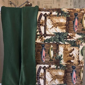 May include: A green shower curtain and a patterned curtain with a wilderness theme. The patterned curtain features images of fish, deer, and a bear, set against a backdrop of wood and pine needles. The text "Wilderness" is visible.