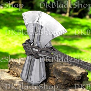 May include: A silver and gray battle axe with a wooden handle. The axe head has a geometric design and is wrapped with a dark brown, vine-like material. The handle is dark gray and the axe is resting on a wooden surface.