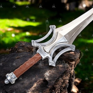 Może przedstawiać: Srebrny i brązowy miecz z szerokim ostrzem i skomplikowaną osłoną. Rękojeść jest owinięta brązową skórą. Miecz spoczywa na ciemnej, teksturowanej powierzchni z rozmytym zielonym tłem.