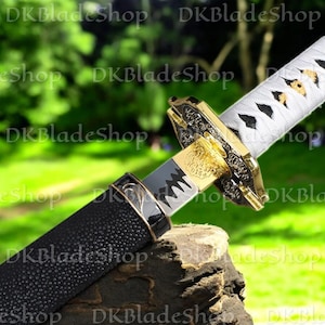 May include: A Japanese katana sword with a black textured handle and a gold-colored guard. The blade is silver and the handle is wrapped in white fabric with black accents. The sword is displayed outdoors with a blurred green background.