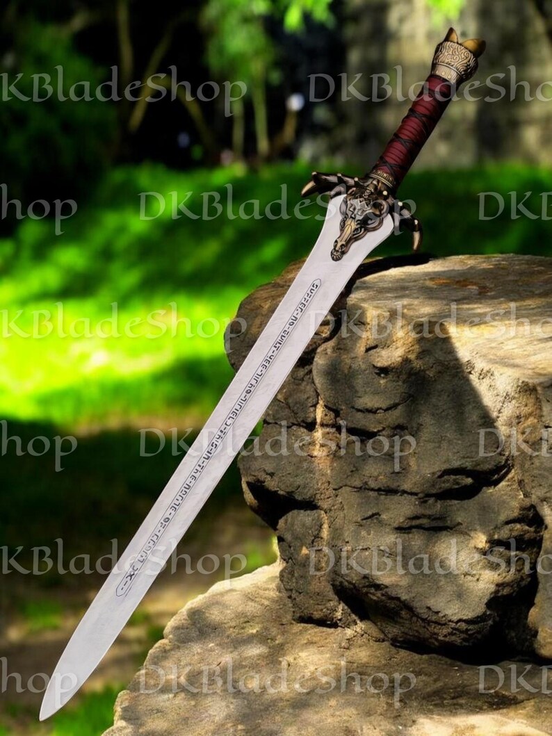 K&ouml;nnte beinhalten: Ein detailliertes Fantasy-Schwert mit einer silbernen Klinge und einem verzierten Bronzegriff. Der Griff ist mit einem dunkelroten Material umwickelt. Die Klinge hat ge&auml;tzten Text. Das Schwert liegt auf einem Felsen. Das Design deutet auf eine Fantasy-Waffe hin.