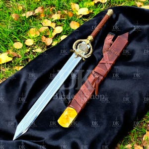 Puede incluir: Una espada decorativa con una hoja plateada y una empuñadura dorada con un diseño ornamentado. El mango está envuelto en cuero marrón y descansa junto a una vaina de cuero a juego. La espada se exhibe sobre una tela negra sobre hierba.