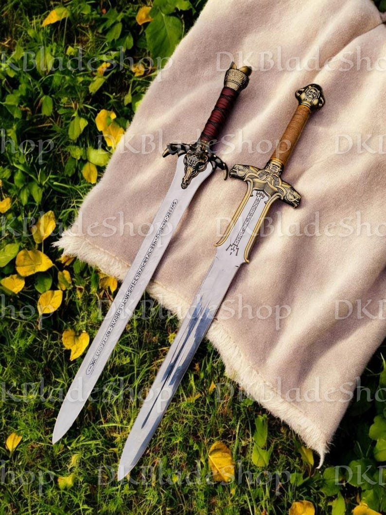 K&ouml;nnte beinhalten: Zwei dekorative Schwerter mit verzierten Griffen und silbernen Klingen liegen auf einem beigen Tuch. Ein Schwert hat einen rotbraunen Griff, das andere einen beigen Griff. Die Klingen sind mit Text und dekorativen Mustern graviert.