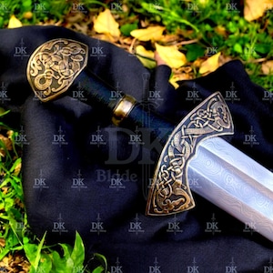 May include: A detailed image of a sword with a bronze-colored hilt and pommel featuring intricate Celtic knot designs. The blade has a silver finish. The sword rests on a black cloth, with green foliage in the background.