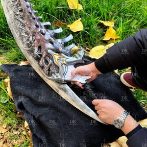 May include: A large, ornate sword with a silver blade and a detailed hilt featuring gold and bronze accents. The hilt has a textured, organic design. The sword is held against a black cloth and green grass background.