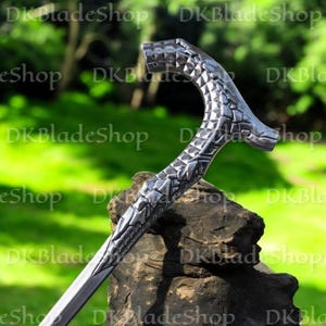 May include: A silver walking cane featuring a dragon scale design on the handle and shaft. The handle is curved and shaped like a dragon's head. The cane is displayed on a dark, textured surface with a blurred green backdrop.