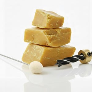 Puede incluir: Una pila de tres bloques de cera de abeja de color amarillo dorado, una broca metálica con una espiral negra y plateada, una herramienta metálica con un lazo y una pequeña bola blanca. Sobre una superficie blanca.