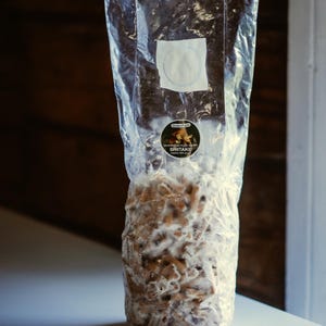 以下が含まれることがあります： シイタケ栽培培地が入った透明なビニール袋。袋には白いラベルと「GreenFarm Mushroom Plus Shiitake」と書かれた丸いステッカーが付いています。内容は茶色と白の混合です。