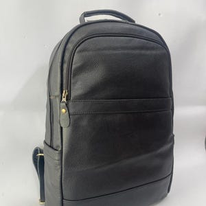 May include: A black backpack with a textured finish, featuring a top handle, adjustable shoulder straps, and a front zippered pocket. The design includes a horizontal seam across the front and a gold-toned metal zipper pull with a leather tab.
