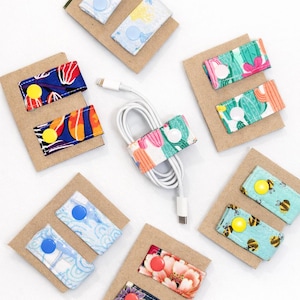 May include: A collection of fabric cable organizers with various patterns and colors. Designs include floral, geometric, and bee motifs. Each organizer is attached to a small cardboard backing and secured with a snap closure. A white charging cable is also shown.