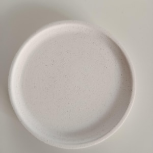 Könnte beinhalten: Ein runder, cremefarbener Keramikteller mit leicht strukturierter Oberfläche. Der Teller hat eine geringe Tiefe und ein schlichtes, minimalistisches Design, das sich zum Servieren von Speisen oder als dekoratives Element eignet. Der Teller befindet sich auf weißem Hintergrund.