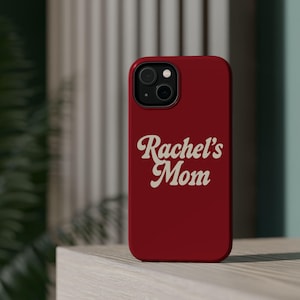 May include: A red phone case with the words "Rachel's Mom" in a white, cursive font. The case has a camera cutout and is sitting on a light-colored surface.