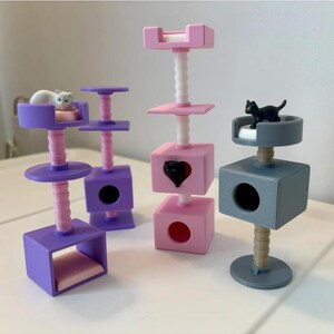 Könnte beinhalten: Vier Miniatur-Kratzbäume in den Farben Lila, Pink und Grau. Zwei lila Bäume haben eine weiße Katzenfigur oben. Ein hoher pinker Baum hat einen weißen Pfosten und eine schwarze Katzenfigur in einem herzförmigen Ausschnitt. Ein grauer Baum hat eine schwarze Katze.