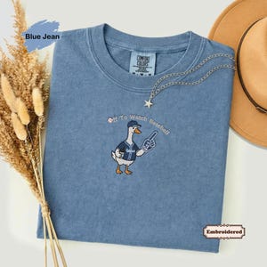 May include: A blue jean colored t-shirt featuring an embroidered graphic of a goose wearing a baseball cap and jersey, holding a foam finger with the text Off To Watch Baseball. The shirt has a Comfort Colors label at the inner neckline.