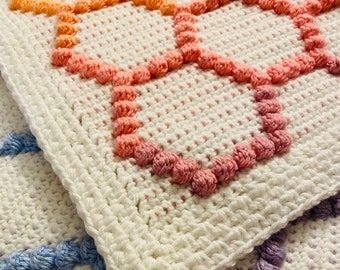 Puede incluir: Primer plano de una manta de crochet blanca con pompones de diferentes colores alrededor de los bordes. Los pompones son en tonos de rosa, azul y morado.