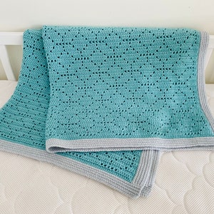 Puede incluir: Una manta de bebé de crochet azul claro con un borde gris. La manta está doblada y puesta sobre un colchón de cuna blanco.