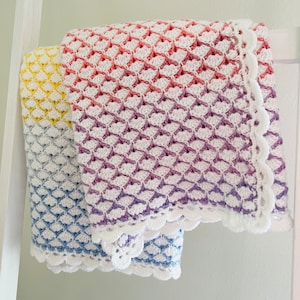 Puede incluir: Tres mantas de bebé de ganchillo en diferentes degradados de color. Las mantas están hechas con hilo blanco y tienen un delicado borde festoneado. Las mantas están apiladas una encima de la otra, con las mantas amarilla y azul visibles debajo de la manta rosa y morada.