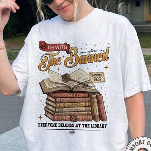 Könnte beinhalten: Weißes T-Shirt mit einer Grafik, die einen Stapel Bücher, einen Schlüssel und den Text "I'M WITH The Banned" zeigt. Das Design enthält die Worte "Banned Book Club" und "EVERYONE BELONGS AT THE LIBRARY."