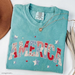 May include: Seafoam green t-shirt with the word "AMERICA" in patchwork letters and star accents. The shirt has a crew neck and a small tag at the neckline. A gold necklace with a pendant is draped across the shirt.