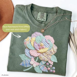 以下が含まれることがあります： オリーブグリーンのTシャツ。フェイクパッチワークの花柄のデザイン。パステルカラーの花びら、葉、蝶が特徴です。ペンダント付きのゴールドネックレスがTシャツにかけられています。「Faux Patchwork Print Effect」の文字が見えます。