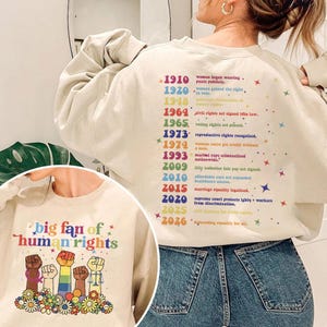 May include: Beige sweatshirt with the text "big fan of human rights" and a graphic of raised fists in various colors. The back of the sweatshirt lists historical dates and events related to women's rights and equality.