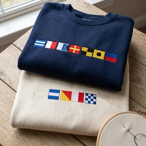May include: Two sweatshirts, one navy blue and one cream, are displayed on a wooden surface. Both sweatshirts feature embroidered nautical signal flags in various colors. The navy sweatshirt is on top, with the cream one below. A needle and embroidery hoop are in the foreground.
