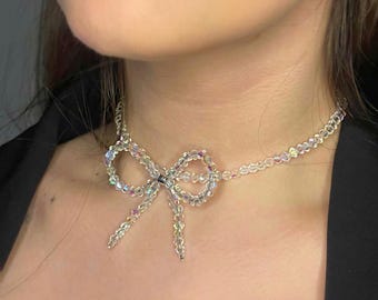 May include: A clear beaded necklace featuring a bow pendant. The bow is crafted from iridescent beads, and the necklace is worn around the neck. The beads reflect multiple colours, creating a shimmering effect. The necklace is a delicate piece of jewellery.