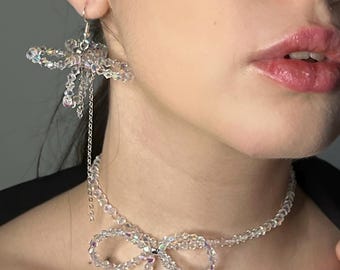 May include: A clear crystal beaded jewellery set featuring a bow-shaped necklace and matching earrings. The earrings have a floral design with dangling chains. The necklace and earrings are made of clear, iridescent beads.