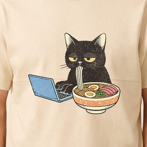 Op de afbeelding: Een crèmekleurig T-shirt met een afbeelding van een zwarte kat die ramen eet en een blauwe laptop gebruikt. De ramenkom bevat noedels, eieren en groenten. Het woord "Natural" staat onderaan het shirt.