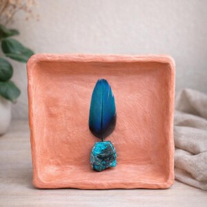 May include: A teal blue feather is displayed on a small, square, terracotta-colored tray. The feather is set on a small, rough, turquoise-colored stone. The tray sits on a light-colored wooden surface.