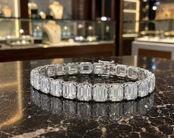 Puede incluir: Una pulsera de tenis en tono plateado con una hilera continua de piedras transparentes de corte esmeralda engastadas en eslabones metálicos. La pulsera está dispuesta en forma circular e incluye un cierre metálico seguro en la parte posterior.