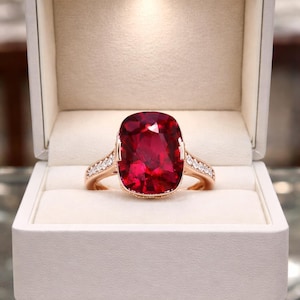 May include: A rose gold ring with a large, cushion-cut, deep red gemstone. The band is adorned with small, clear accent stones. The ring is presented in a white, illuminated jewelry box, creating a luxurious display.