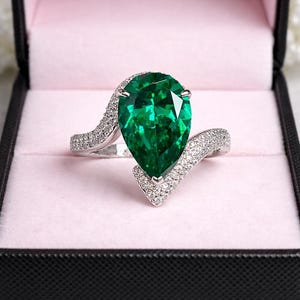 May include: A silver ring with a large, pear-cut emerald gemstone, presented in a black box with a pink lining. The ring's band has a wave design, embellished with small, sparkling stones.