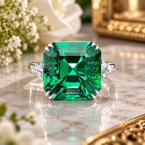 Puede incluir: Un anillo de plata con una gran piedra preciosa de corte cuadrado y color verde vibrante, sujeta por cuatro garras. La banda está acentuada a cada lado por una piedra transparente de corte triangular más pequeña, con acabado metálico.