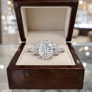 May include: An oval-cut diamond engagement ring with side stones, set in a silver band, displayed in a dark wooden ring box with a cream-colored interior. The ring's large center stone sparkles brightly.